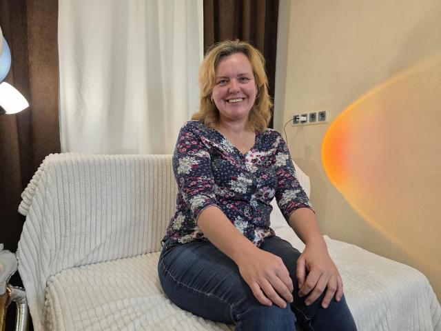 VestaWoody, with blonde hair and a smile, sits on a white, ribbed sofa wearing a floral blouse and blue jeans, hands resting on her knees.