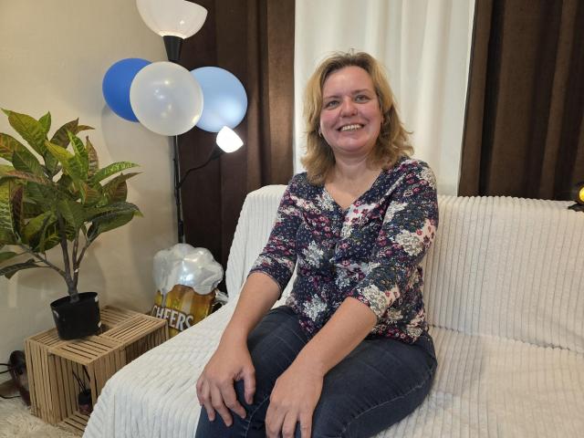 VestaWoody has blonde hair, a floral shirt, and blue jeans, sitting on a white couch, smiling, with balloons and a plant to the left. She's got a cute ass.