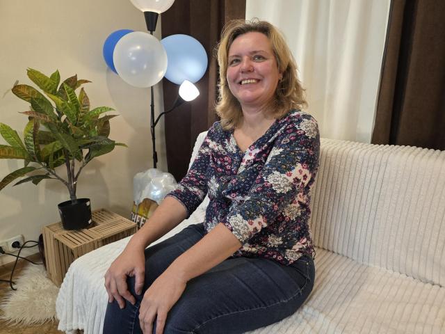 VestaWoody sits on a white couch, wearing a floral shirt and blue jeans, smiling with her hands on her knees, next to a plant and balloons.