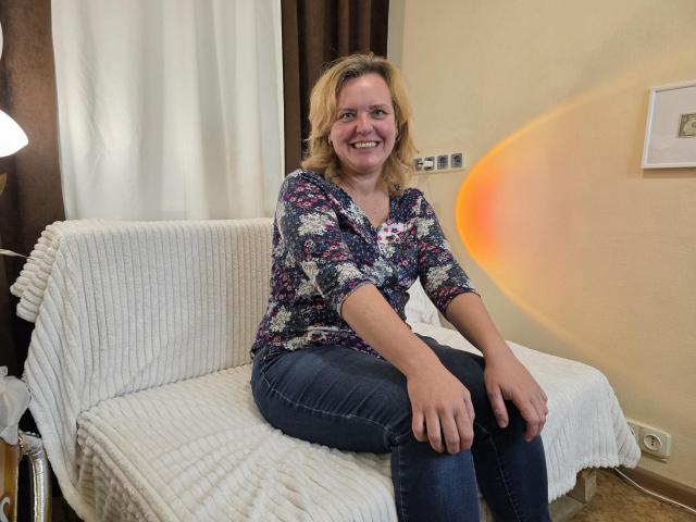 VestaWoody sits on a white, ribbed couch, smiling. She's wearing a floral shirt, blue jeans, and has blonde hair. A warm orange light reflects on the wall.