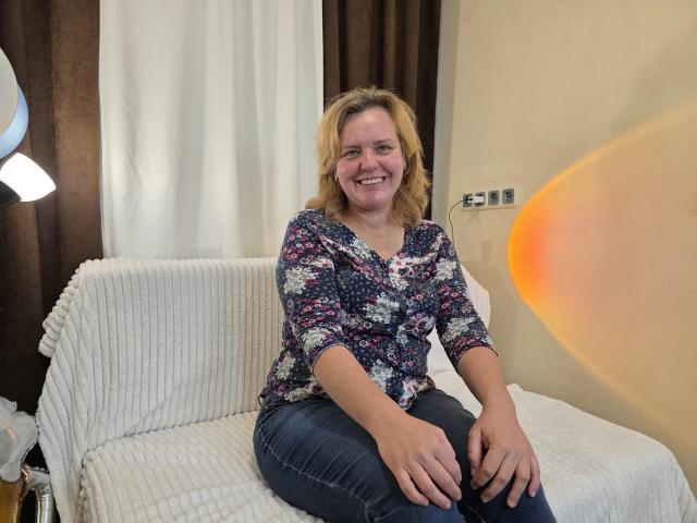 VestaWoody, a blonde woman with medium-sized tits, wearing a floral shirt and blue jeans, sits on a white couch, smiling with hands on her knees.
