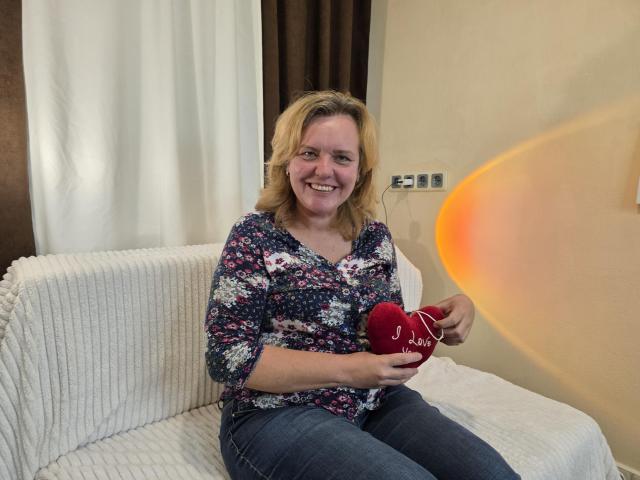 VestaWoody, with blonde hair and a floral shirt, holds a red heart-shaped mug, smiling on a white couch, her ass barely visible under her jeans.