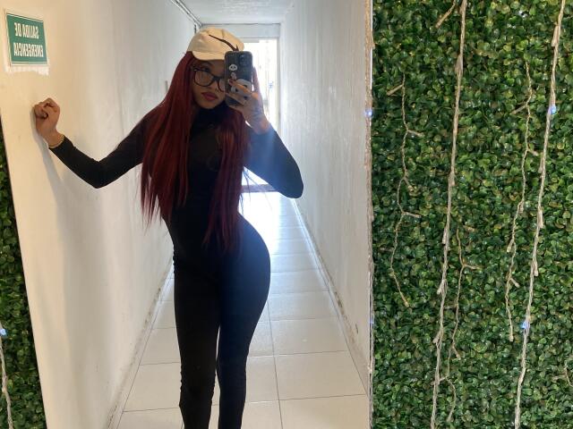 KathaThoms with long red hair, black long-sleeve bodysuit, glasses, white cap, holding phone in mirror selfie, showing curvy ass and small tits, standing in white hallway.