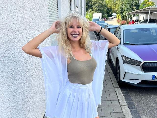 BabsiMonroe-hot with blonde hair, smiling, wearing a sheer white robe over a green tank top and white skirt, standing by a purple and white car.