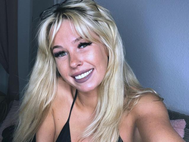 BabsiMonroe - atraente, com cabelo loiro, olhos azuis e um grande sorriso, vestindo um top preto de alças, mostrando seus seios, e com um rosto bonito e sexy..