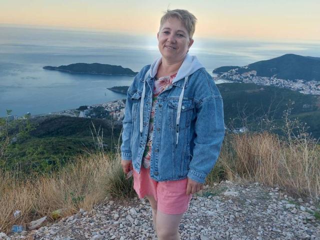 LunaRossaa stands with a smile, wearing a denim jacket, white hoodie, pink shorts, and a floral shirt, on a rocky hilltop with grass, at sunset, near a coastal town.