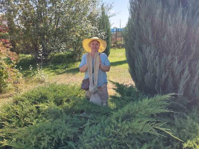 LunaRossaa standing in a grassy yard, wearing a yellow hat, blue shirt, beige pants, and a brown purse, with greenery and a tall tree in the background.