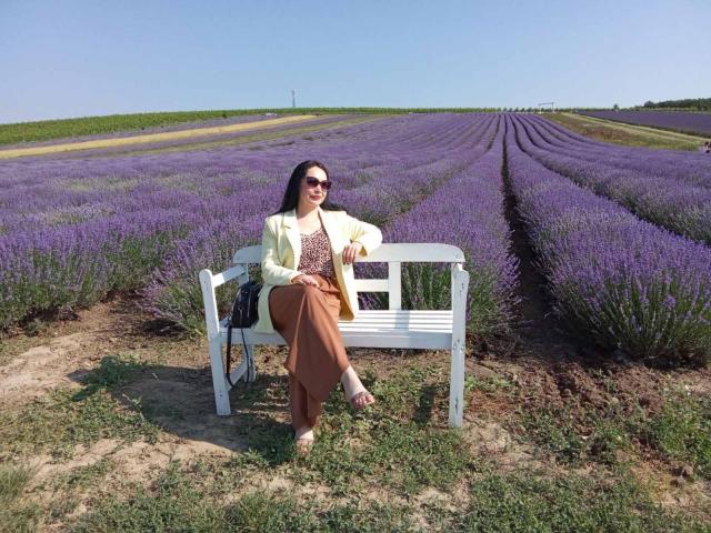 JosefinaWelpo, with purple hair, sunglasses, yellow cardigan, brown pants, and sandals, sits on a white bench in a lavender field, holding a drink, with a bag on the bench.