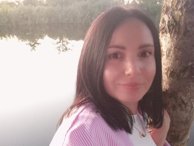 JosefinaWelpo has medium-length black hair, a pink striped shirt, and a necklace with a small pendant, smiling softly with a slight blush on her face.