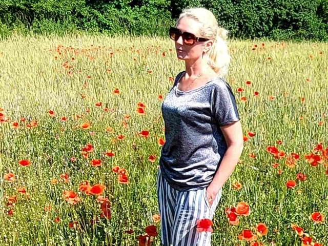 EmmaParkk with blonde hair, wearing sunglasses, gray tee, and striped pants, standing in a field of red poppies, hands in pockets, sunny day, green trees in background.