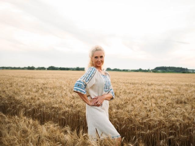 EmmaParkk stands in a golden wheat field, wearing a white dress with blue embroidered patterns on the sleeves, smiling with hands on her hips, blonde hair in a ponytail.