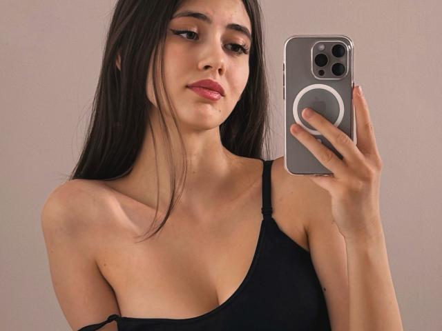 SweetyVikky with long dark hair, wearing a black spaghetti strap top, holding a phone with a ring grip, showing her bare shoulders and small boobs, and a slight smile.