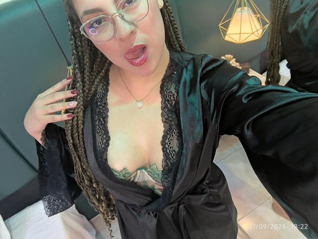 VictoriaKurt with braided hair, glasses, and a black lace robe, showing her tits with a green tattoo under her cleavage, tongue out, and red nails. (Selfie, date visible)