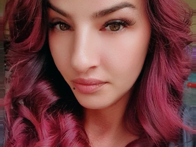 TonPlaisirDeReverBb has pink curly hair, brown eyes with long lashes, and a small beauty mark on her lower lip. Her skin is smooth with a slight pinkish tone.