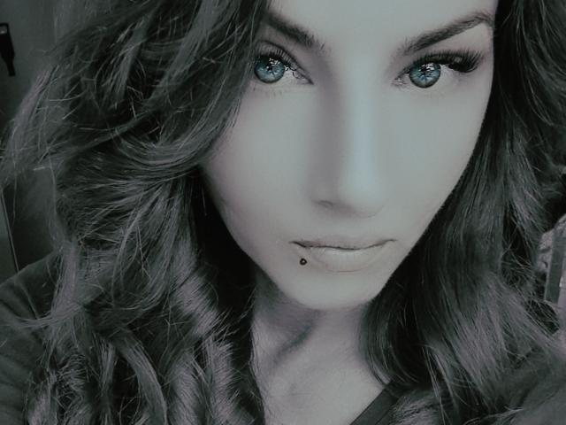 TonPlaisirDeReverBb's face with wavy grey-blue hair, piercing blue eyes, long eyelashes, and a small black beauty mark below her lip, wearing a dark shirt.