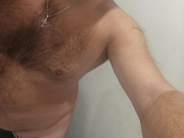 EffectGerald's hairy chest with small nipples, wearing a silver chain necklace, has a tattoo on his right shoulder. His arm is slightly raised, showing some body hair.