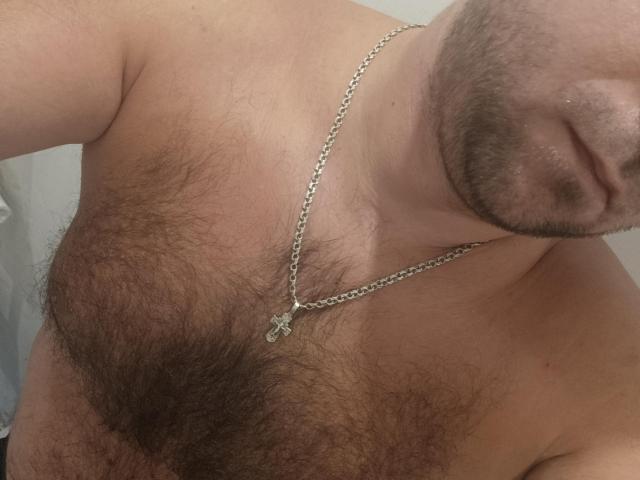 Dê a Gerald um colar de prata com um pequeno crucifixo, destacando seu peito peludo, e um rosto parcialmente visível com barba. Sua boca está ligeiramente aberta, revelando um pouco de seus lábios..