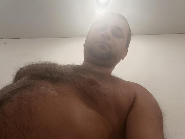 Efeito: Gerald, sem camisa, com o peito peludo e cabelo curto e escuro, olhando de baixo para cima com um brilho suave acima. Sua expressão facial é neutra e ligeiramente cansada..