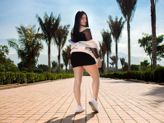 JeniferPowell with long black hair, off-shoulder black top, black mini-skirt, white sneakers, and pink jacket, standing on brick pathway with palm trees, looking back, butt slightly raised.