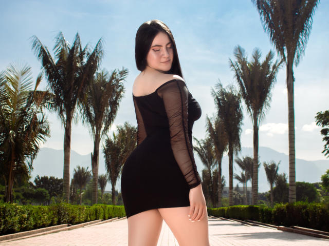 JeniferPowell with long black hair, wearing a sheer black dress, showing her big ass and small tits, standing outdoors, palm trees in background, sunny day.