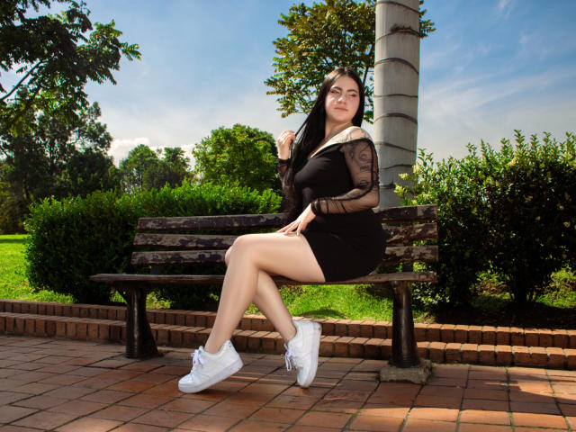 JeniferPowell, with black dress and sheer sleeves, sits on a wooden bench, legs crossed, white sneakers, exposing her bare thighs, dark hair, green eyes, park setting.