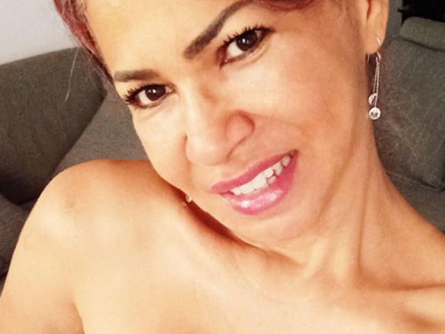 BrigiteWalsh's face, smiling with pink lipstick, brown eyes, and red hair, wearing small hoop earrings, topless with visible shoulder, looking at the camera.