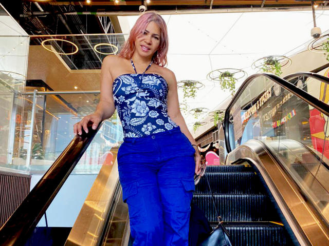 BrigiteWalsh with pink hair, blue floral strapless top, and blue cargo pants, standing on an escalator, smiling, holding her ass with her hand, looking sexy.