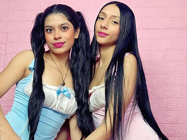 HarleyAntella with long black hair, pink lipstick, and small tits in blue and pink dresses, standing side-by-side, pink brick wall background, smiling at camera, arms around each other.