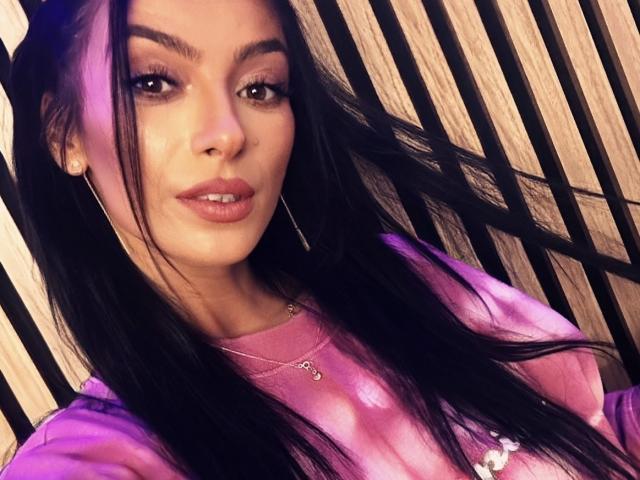 KinkyAmelie, con i suoi lunghi capelli neri, la maglietta rosa tie-dye e gli orecchini a cerchio, mostra le sue labbra rosa carnose e il trucco leggero. Il suo seno è piccolo e la sua vulva è invisibile..