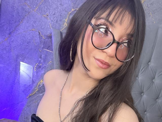 ValuuRodriguez with glasses, pink blush, and pink lipstick, has long dark hair, a black off-shoulder top, and a necklace, looking at the camera with a slight smile.