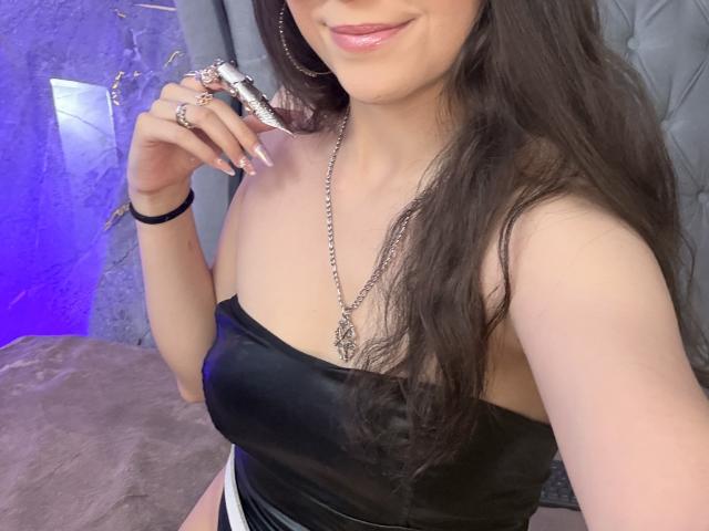 ValuuRodriguez with long brown hair, pink lipstick, black leather strap dress, silver chain necklace, holding a silver ring, showing off her tits, and a black bracelet.