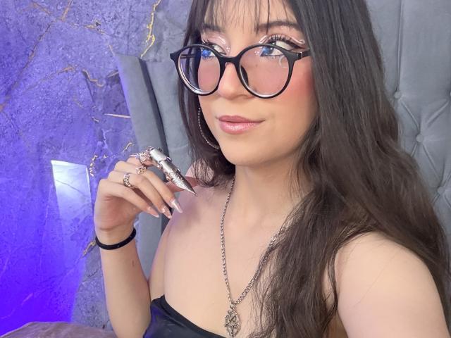 ValuuRodriguez with long black hair, pink glossy lips, black glasses, pink eyeshadow, wearing a black strapless top, holding a silver ring, smiling, showing off pink nail polish.