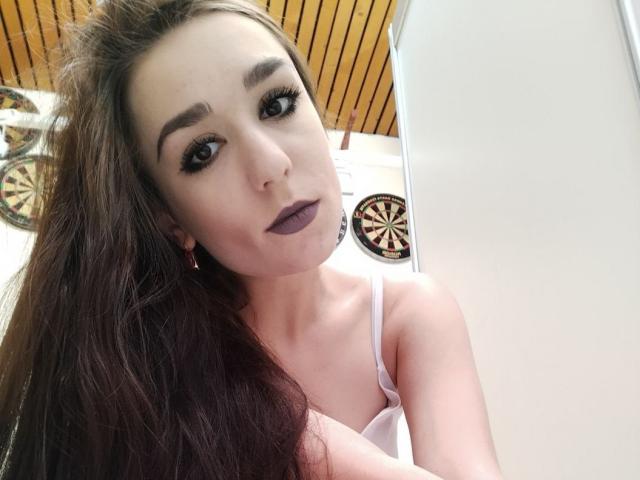 LilithNoir with long brown hair, dark purple lipstick, and white tank top, showing her tits, looking at the camera with black eyeliner and pink earrings.