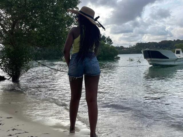 Africanah stands on a beach with her back to the camera, wearing a straw hat, yellow top, and frayed denim shorts, holding a fishing line, by a boat.