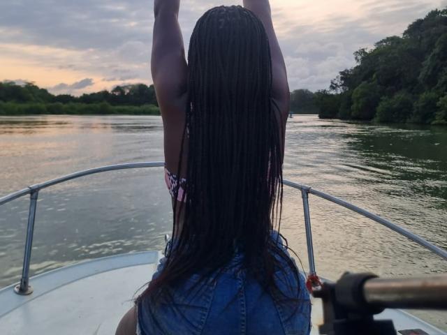Africanah with long braided hair, arms raised, wearing a denim vest, sitting on a boat, water in front, sunset, camera in bottom right, river on the right.