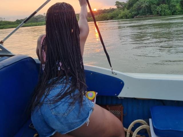 Eine afrikanische Frau mit langen Zöpfen, blauen Jeansshorts und einem pinken Oberteil, die einen Stock hält und in einem blauen Boot sitzt, mit dem Rücken zur Kamera, während der Sonnenuntergang sich im Wasser spiegelt..