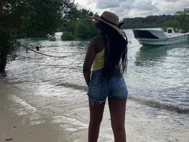 Africanah stands on a beach, facing water, wearing a straw hat, yellow tank top, and blue denim shorts, with long black dreadlocks, and a visible ass. Boat in background.