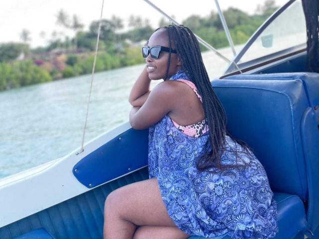 Africanah with dark skin, long braids, wearing a blue patterned dress and black sunglasses, sitting on a blue boat, smiling, showing her ass, with her hand on her chin.
