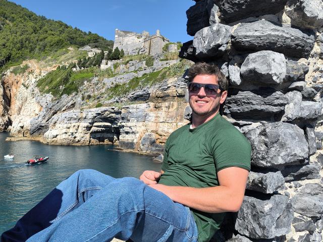 LavForYou, a muscular dude with short brown hair, green t-shirt, and blue jeans, sits against rocky cliffs, wearing dark sunglasses, with a boat and castle in the background.