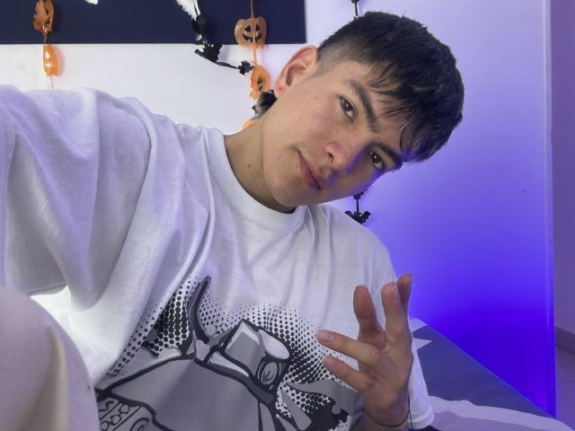 Archangel69, com cabelo preto curto, camisa branca com estampa, com um leve sorriso, levantando um dedo, em um quarto com decorações de Halloween em tons de roxo e laranja..