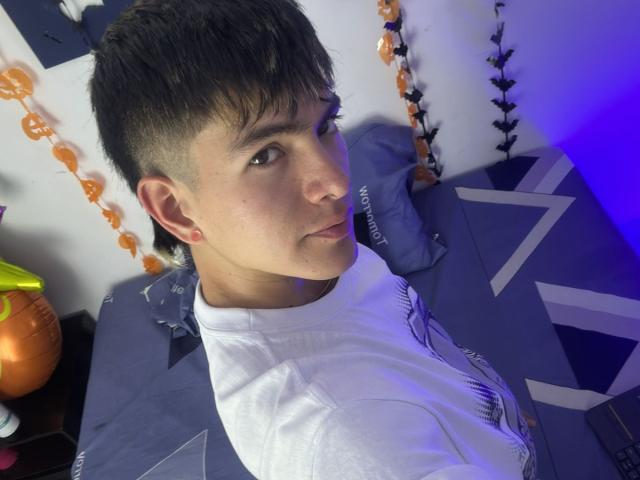 Archangel69 heeft kort donker haar, kleine roze oorbellen en een wit shirt. Hij kijkt over zijn schouder met een lichte glimlach, waarbij zijn neus en een stukje van zijn achterwerk zichtbaar zijn..