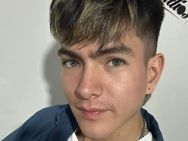 Archangel69 a un visage mignon, légèrement brillant, avec des yeux verts, des cheveux bruns et blonds, un petit piercing au nez, et porte un sweat à capuche bleu avec un t-shirt blanc..