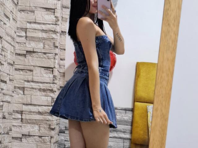 LizzeJoy with black hair, wearing a blue denim dress, showing her ass, taking a mirror selfie, her hand tattooed, in a room with stone and yellow walls.