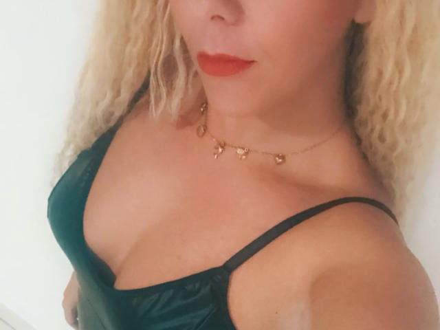 Osmoze has blonde curly hair, red lipstick, and wears a green satin top with black straps. She has a gold necklace with small heart charms. Her tits are visible.