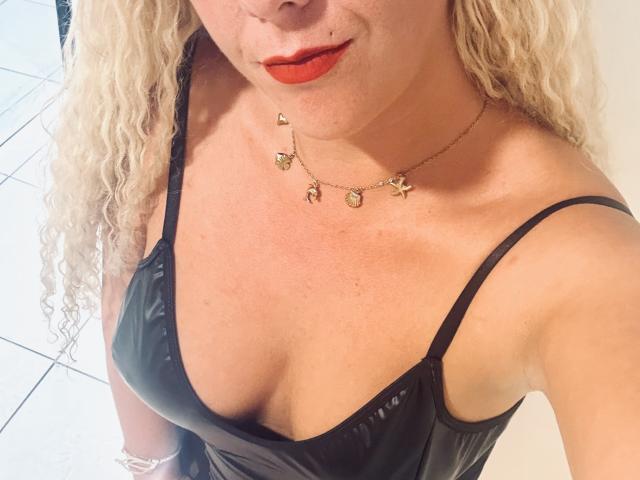 Osmoze with curly blonde hair, wearing a black shiny dress with a low neckline, showing her tits, red lipstick, gold necklace with small charms, and a white tiled floor.