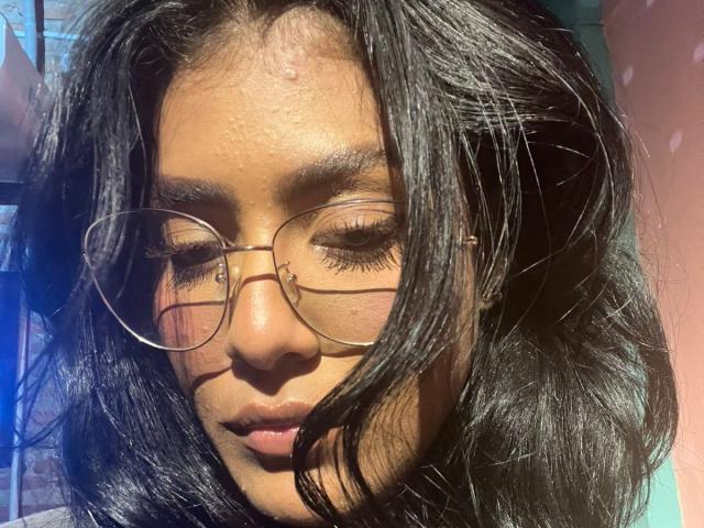Babilett, com cabelos escuros e ondulados, pele morena e nariz pequeno, usando óculos redondos, olhos fechados, a luz do sol realçando seu rosto e um leve sorriso em seus lábios rosados..