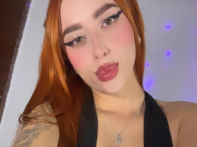 SarahLuxe with red hair, black eyeliner, pink cheeks, and glossy pink lips, wearing a black halter top, showing a tattoo on her left shoulder.