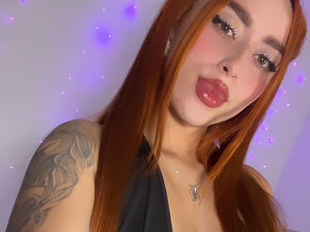 SarahLuxe with red hair, pouty lips, tattoo on right shoulder, black halter top, necklace, and subtle makeup, looking at camera with slightly parted pink lips.