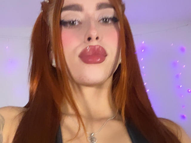 SarahLuxe with red hair, puffy pink lips, and a black top, making a pouty face, wearing a silver necklace, her eyes half-closed, looking cute and sexy.