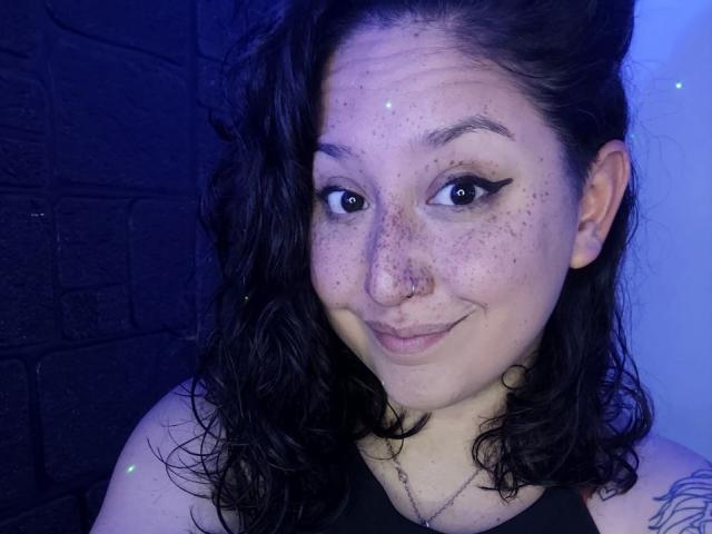 SweetYamii with dark wavy hair, freckles, nose ring, and black eyeliner, smiling gently, blue-lit, showing tattoo on right shoulder, wearing black top, under soft blue light.